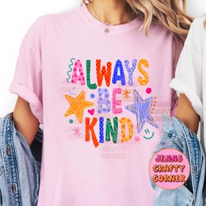 May include: A light pink t-shirt with the words "ALWAYS BE KIND" in colorful, playful lettering. The design includes stars, hearts, and a smiley face. The shirt is a casual, everyday wear item.