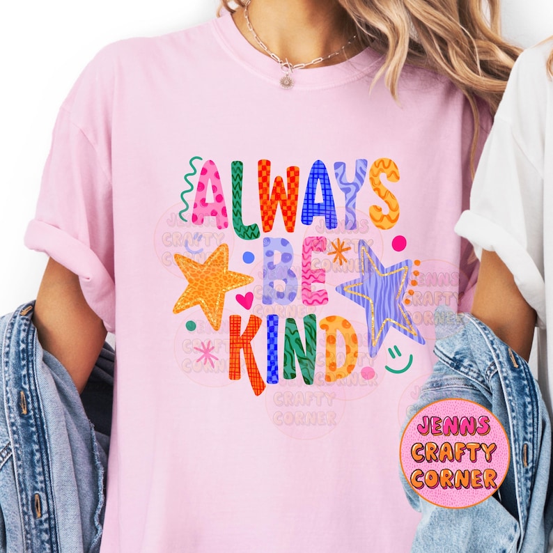 May include: A light pink t-shirt with the words "ALWAYS BE KIND" in colorful, playful lettering. The design includes stars, hearts, and a smiley face. The shirt is a casual, everyday wear item.