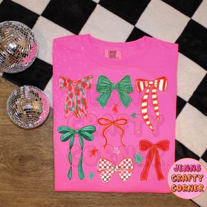 May include: A pink t-shirt featuring various bow designs in red, green, and white. The bows have different patterns and styles, with stars and the text "JENNS CRAFTY CORNER" on the bottom right. Two disco balls are on the left.