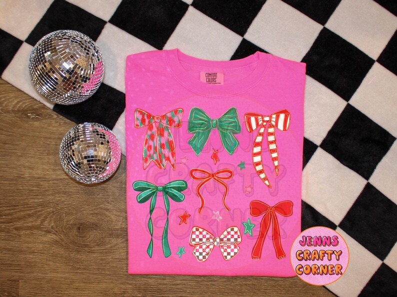 May include: A pink t-shirt featuring various bow designs in red, green, and white. The bows have different patterns and styles, with stars and the text "JENNS CRAFTY CORNER" on the bottom right. Two disco balls are on the left.