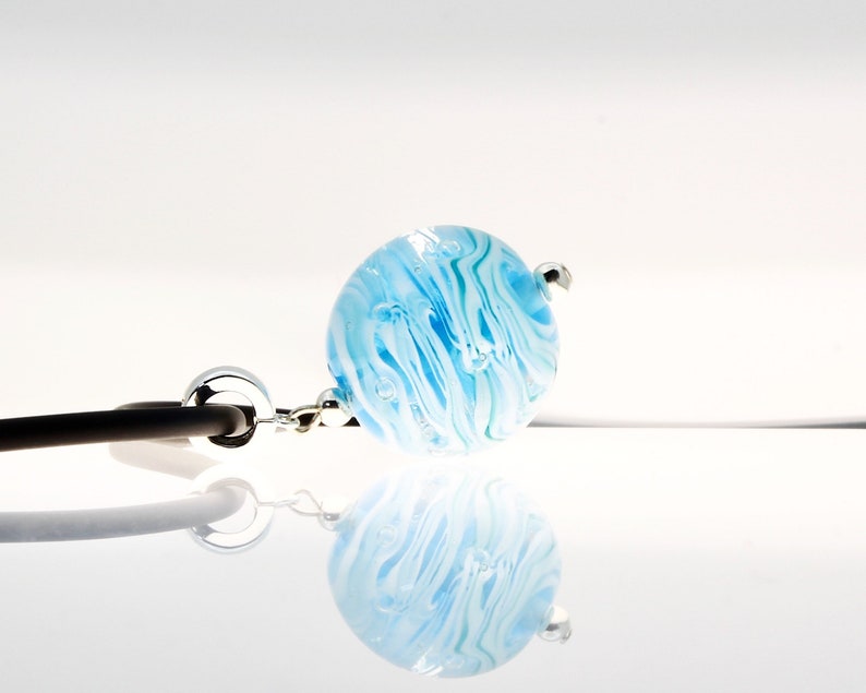 Puede incluir: Un orbe de vidrio azul y blanco con una cadena plateada unida. El orbe tiene un patr&oacute;n de remolino y parece tener burbujas de aire atrapadas en su interior.