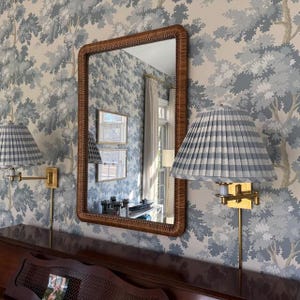 May include: A rectangular mirror with a brown woven rattan frame is mounted on a wall covered in blue and cream tree-patterned wallpaper. Two brass wall sconces with pleated blue and white gingham fabric shades flank the mirror.