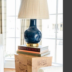 May include: A navy blue lamp with a woven beige shade sits atop a stack of books on a wooden side table. The lamp has a gold base and a spherical body. The books include "RALPH LAUREN" and "PORTRAITS OF HOME".