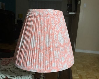 Puede incluir: Pantalla de lámpara plisada rosa con un estampado floral blanco. La pantalla tiene forma de cono y está sobre una base de lámpara. La lámpara está sobre una mesa de madera.