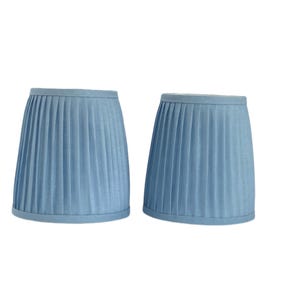 May include: Two light blue pleated lampshades. The lampshades have a classic, cone-like shape with vertical pleats. The fabric appears to be a soft, solid color, and the shades are designed to fit on a lamp base. The lampshades are a light blue color.