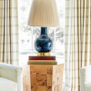 May include: A navy blue lamp with a woven shade sits atop a wooden side table. The lamp is placed in front of a window with cream-colored curtains. A checkered ottoman is in the foreground, and two white armchairs are on either side.