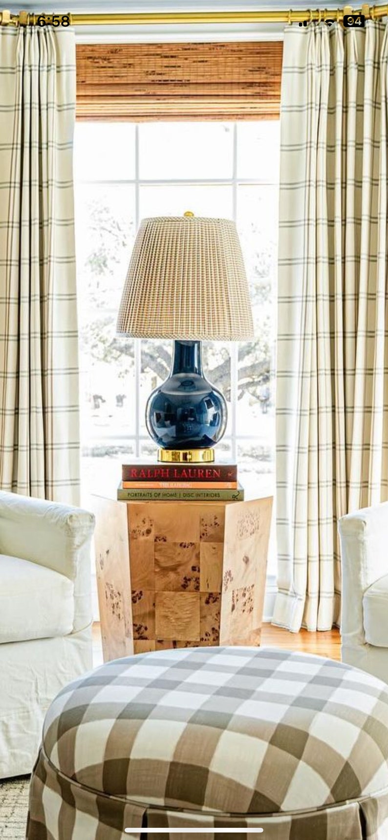 May include: A navy blue lamp with a woven shade sits atop a wooden side table. The lamp is placed in front of a window with cream-colored curtains. A checkered ottoman is in the foreground, and two white armchairs are on either side.