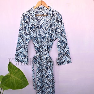 May include: A white and blue patterned robe with a long sleeve and a tie belt. The robe is hanging on a wooden hanger against a light pink wall.