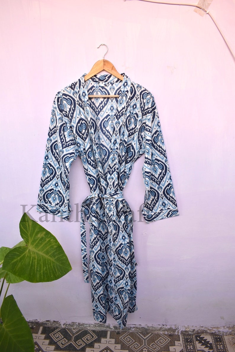 May include: A white and blue patterned robe with a long sleeve and a tie belt. The robe is hanging on a wooden hanger against a light pink wall.