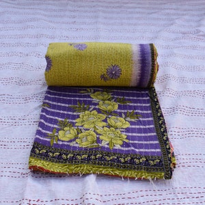 Op de afbeelding: Een opgerolde, olijfgroene en paarse bloemenquilt met een bijpassend uitgevouwen gedeelte. De quilt heeft een groot bloemmotief in geel en groen op een paarse gestreepte achtergrond. De randen zijn afgewerkt met een donkergroene en zwarte rand.