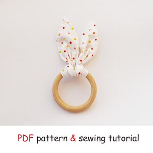 May include: A wooden teething ring with a white fabric bunny ear attached. The fabric has red, yellow, and orange polka dots.