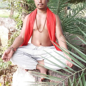 May include: A man sits in a meditative pose with his legs crossed, wearing a red shawl and a beaded necklace. He is surrounded by lush green foliage.