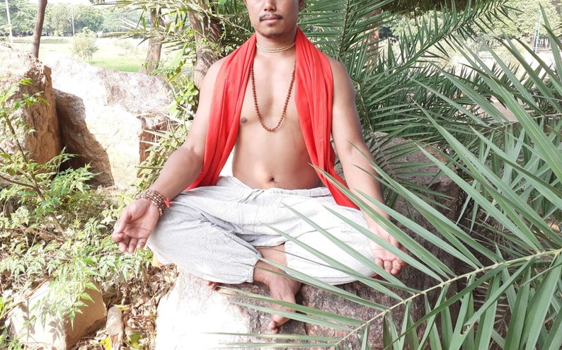 May include: A man sits in a meditative pose with his legs crossed, wearing a red shawl and a beaded necklace. He is surrounded by lush green foliage.