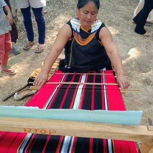 May include: A woman in a black dress and beaded necklace is weaving a red, black, and white striped fabric on a wooden loom. The fabric is being woven with a traditional technique.