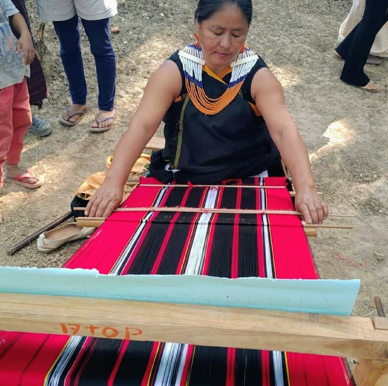 May include: A woman in a black dress and beaded necklace is weaving a red, black, and white striped fabric on a wooden loom. The fabric is being woven with a traditional technique.
