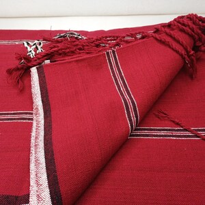May include: A folded red textile with white and black stripes and fringe. The fabric is a solid red color with a textured appearance. The stripes are near the edge of the textile. The fringe is a twisted red cord.