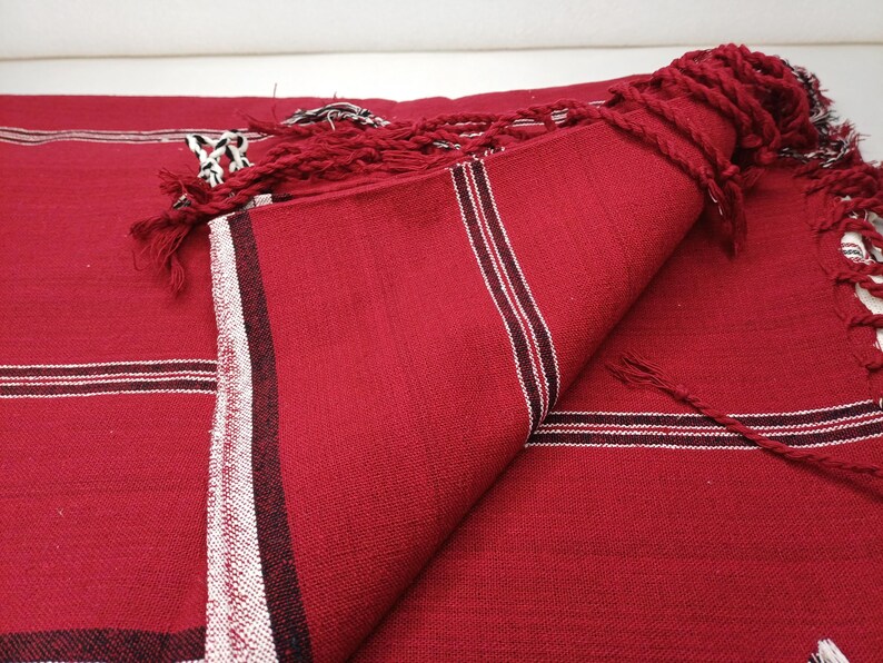 May include: A folded red textile with white and black stripes and fringe. The fabric is a solid red color with a textured appearance. The stripes are near the edge of the textile. The fringe is a twisted red cord.