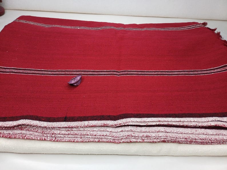 May include: A folded red textile with horizontal stripes in white and dark red. A small, purple decorative element rests on the textile. The textile is stacked on top of a white textile.