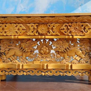 May include: A wooden table with intricate carvings of mythical creatures and floral designs. The table is made of light brown wood and has a rectangular shape.