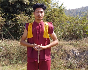 May include: A young man wearing a maroon and yellow sleeveless robe with a long string in his hands. He is standing in front of a green grassy area with trees in the background.