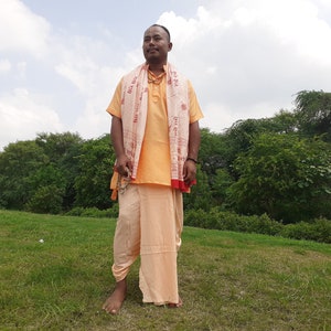May include: A man wearing a light orange shirt and pants with a white and red patterned scarf draped around his shoulders. He is standing in a grassy field with trees in the background.