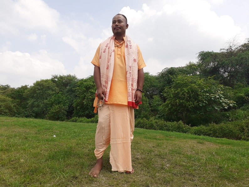 May include: A man wearing a light orange shirt and pants with a white and red patterned scarf draped around his shoulders. He is standing in a grassy field with trees in the background.