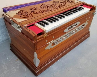 May include: A wooden harmonium with a carved wooden top, a keyboard with white and black keys, and a row of silver knobs. The harmonium is brown with a purple fabric lining the top.