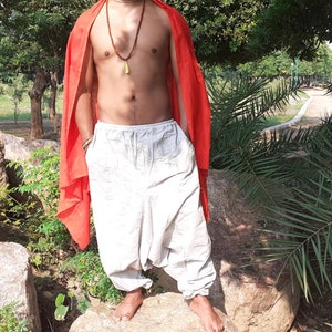 May include: A person wearing white loose-fitting trousers with a red scarf draped over their shoulders. The person is standing on a large rock in a park setting.