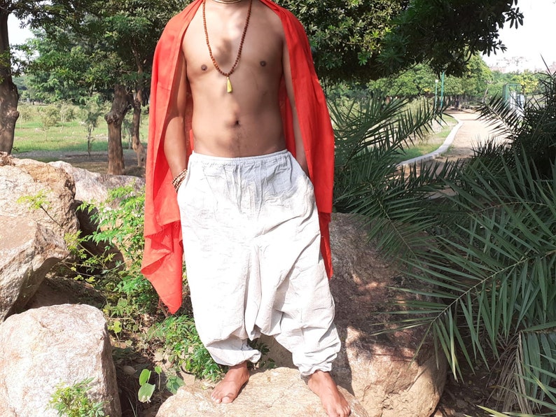 May include: A person wearing white loose-fitting trousers with a red scarf draped over their shoulders. The person is standing on a large rock in a park setting.