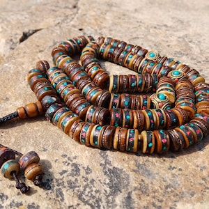 May include: A brown wooden mala prayer bead necklace with turquoise and red accents. The beads are intricately carved and feature a decorative pattern.