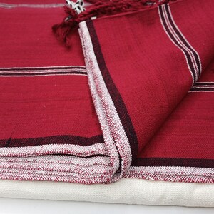 May include: Close-up of a folded red textile with white and black stripes. The edges are finished with a white and red patterned border and red fringe. The textile is layered over a white fabric.