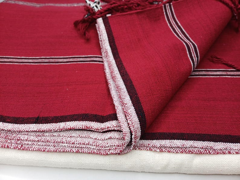 May include: Close-up of a folded red textile with white and black stripes. The edges are finished with a white and red patterned border and red fringe. The textile is layered over a white fabric.