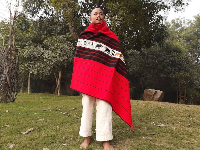 May include: A person wearing a red and black woven shawl with a white stripe and animal designs. The shawl is draped over the person's shoulders and is worn with white pants.
