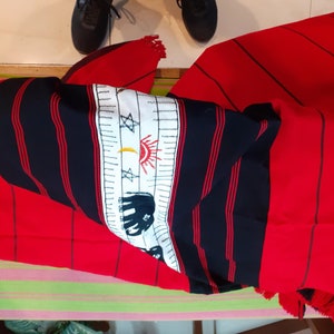 May include: Red and black striped fabric with white embroidered designs, including a sun, moon, stars, and an elephant. The fabric has fringe on the edges.