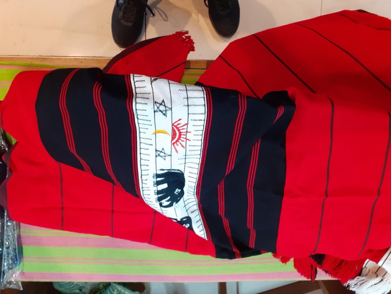 May include: Red and black striped fabric with white embroidered designs, including a sun, moon, stars, and an elephant. The fabric has fringe on the edges.