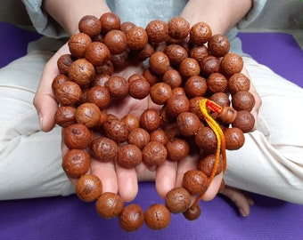 May include: A brown wooden mala bead necklace with a yellow and orange string. The necklace is held in the hands of a person.