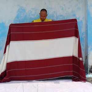 May include: A large, rectangular textile with a striped pattern in burgundy and white. The textile is being held up, showcasing its design. The background is a blue and white wall.
