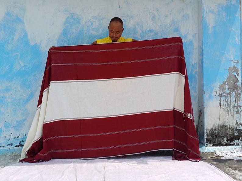 May include: A large, rectangular textile with a striped pattern in burgundy and white. The textile is being held up, showcasing its design. The background is a blue and white wall.