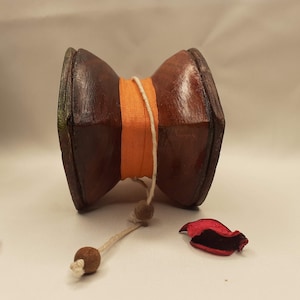 Puede incluir: Un molinillo de oración de madera con una banda de rayas marrones y naranjas y una cuerda blanca con cuentas marrones. El molinillo de oración está sobre una superficie blanca.