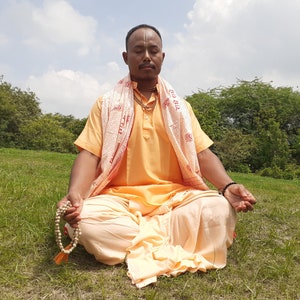 May include: A man in a yellow and white robe sits cross-legged on a grassy field with his eyes closed. He is holding a string of wooden beads in his hands.