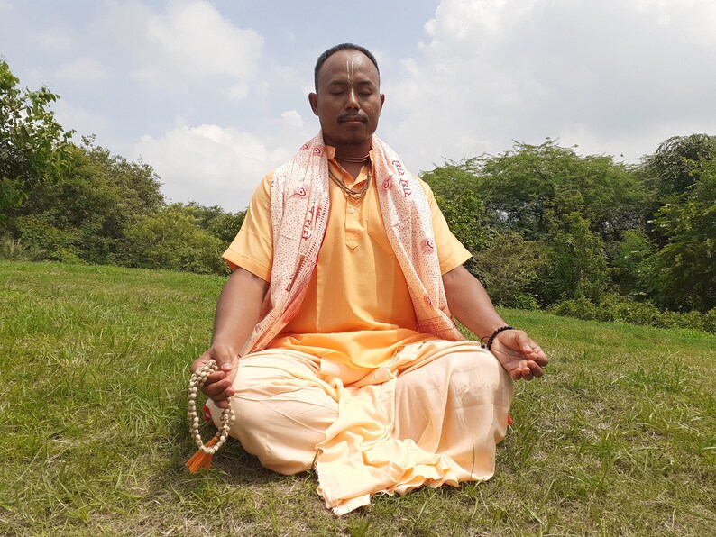 May include: A man in a yellow and white robe sits cross-legged on a grassy field with his eyes closed. He is holding a string of wooden beads in his hands.