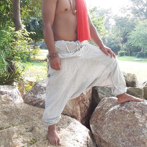 May include: A person wearing loose-fitting, white, linen trousers with a red scarf around their waist. The trousers have a drawstring waist and are gathered at the ankles. The person is standing on rocks in a natural setting.