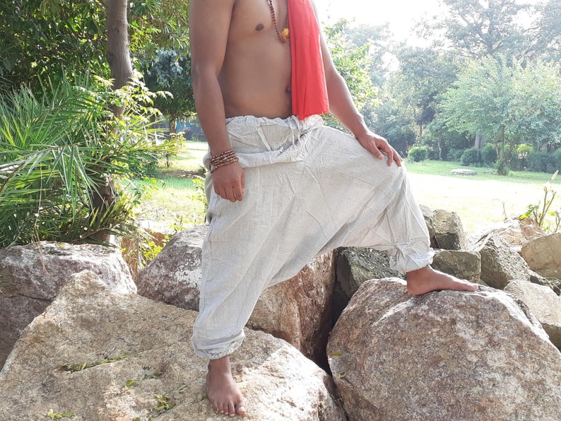 May include: A person wearing loose-fitting, white, linen trousers with a red scarf around their waist. The trousers have a drawstring waist and are gathered at the ankles. The person is standing on rocks in a natural setting.