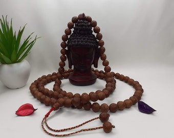 May include: A brown wooden mala bead necklace with a red and white string tassel. The necklace is draped around a small wooden statue of the head of Buddha.