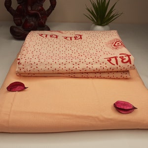 May include: Two folded peach-colored fabric panels with a white fabric panel with red text and a red peacock design on top. The text reads "राधे राधे राधे".