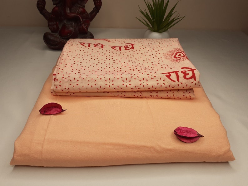 May include: Two folded peach-colored fabric panels with a white fabric panel with red text and a red peacock design on top. The text reads "राधे राधे राधे".