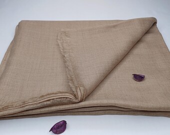 May include: A folded, beige-coloured shawl or blanket with a fringed edge. The fabric has a subtle woven texture. Two small, purple decorative elements are placed on the surface. The item is likely made of a soft, lightweight material.