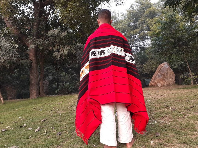 May include: A person wearing a red and black woven shawl with a pattern of elephants and tigers. The shawl has a fringe border and is draped over the person's shoulders.