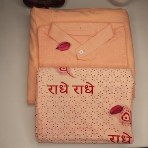 May include: A set of three peach-colored fabric items. The top item is a shirt with a button-up collar. The middle item is a plain fabric rectangle. The bottom item is a patterned fabric rectangle with the text "राधे राधे" in red.