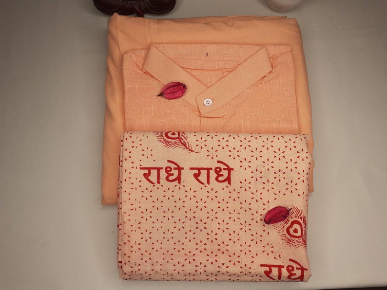 May include: A set of three peach-colored fabric items. The top item is a shirt with a button-up collar. The middle item is a plain fabric rectangle. The bottom item is a patterned fabric rectangle with the text "राधे राधे" in red.